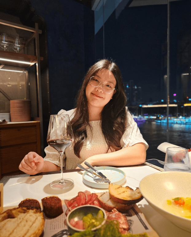Jummum Home Poster_Resized A woman with glasses smiles at the camera while sitting at a restaurant table with a glass of wine and food, with Bangkok and Chao Phraya river in the background at night.